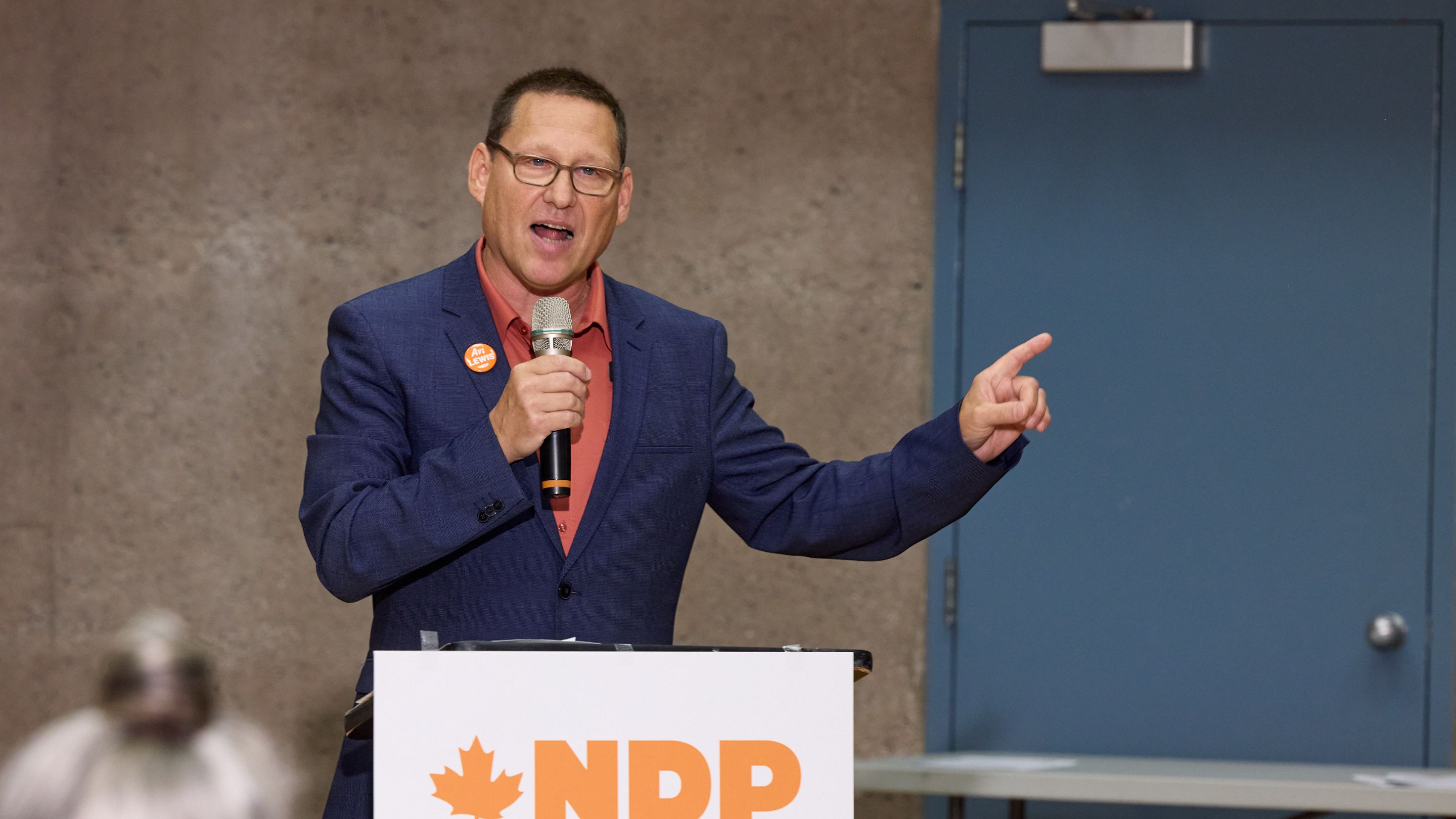 Avi Lewis speaking at an NDP event, gesturing while holding a microphone at a podium